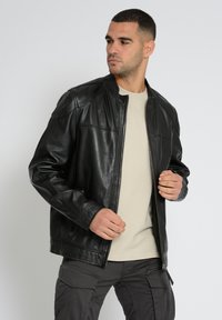 Schwarze Lederjacke mit Reißverschluss, Stehkragen und seitlichen Taschen. Glatte Textur mit gesteppten Details, kombiniert mit einem hellbeigen Hemd.