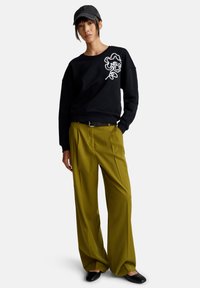 Felpa nera con design floreale bianco, abbinata a pantaloni verdi oliva plissettati e scarpe slip-on nere. Un cappello completa l'outfit.