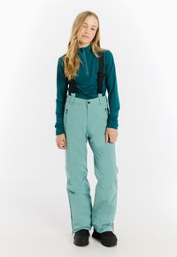 Miękka, tealowa warstwa podstawowa z half-zip i długimi rękawami, w połączeniu z jasnoniebieskimi spodniami narciarskimi z regulowanymi szelkami i kieszeniami. Czarne buty.