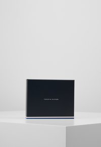 Schwarze rechteckige Box mit mattem Finish, versehen mit dem "TOMMY HILFIGER"-Logo in Weiß und Rot. Ein schmaler blauer und roter Akzent zieht sich entlang des Bodens.