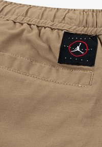 Beige katoenen shorts met een elastische tailleband, voorzien van een zwart label met een rode cirkel en een witte silhouetlogo, en een achterzak.