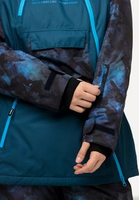 Outerwear turchese con un motivo astratto scuro, caratterizzato da accenti blu, tasche con cerniera e polsini regolabili. Il materiale appare liscio e strutturato.