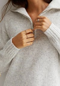 Hellgrauer, gestrickter Pullover mit einem Halbzip-Kragen. Gerippte Bündchen und eine weiche Textur. Hände, die den Reißverschluss verstellen, und silberne Ringe tragend.