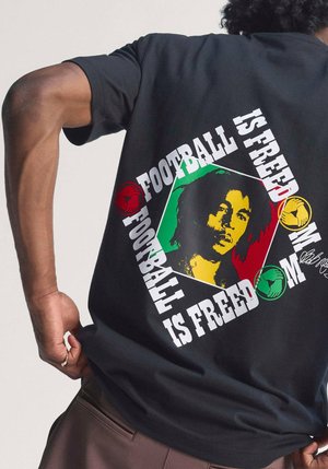 Person wearing a black shirt with a colorful Bob Marley graphic and the text "Football is Freedom" surrounding it on the back.