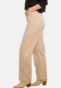 Šviesiai rudos velveto kelnės su tiesia džinsų forma, turinčios nugarines kišenes ir subtilų vertikalių briaunelių tekstūrą. Aprengtos su aukštakulniais batais.