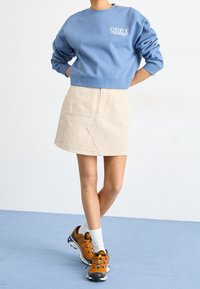 Blå kort sweatshirt med vit "OBEY" logotyp, beige A-linjeformad kjol med sidofickor, och orangea träningsskor med svarta detaljer.