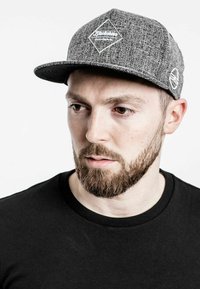 Szürke snapback sapka texturált anyagból, gyémánt logóval és kontrasztos hímzett részlettel az oldalon. Fekete ing alatta.