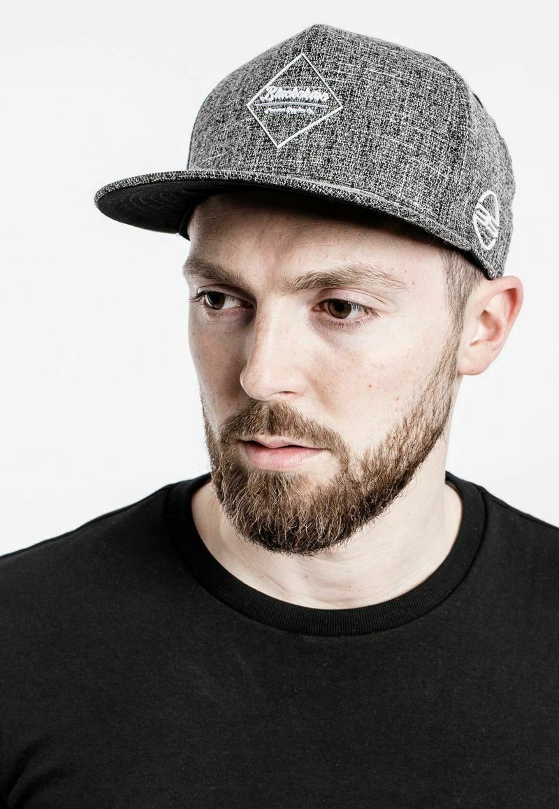 Szürke snapback sapka texturált anyagból, gyémánt logóval és kontrasztos hímzett részlettel az oldalon. Fekete ing alatta.