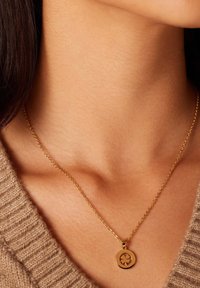 Gouden ketting met een ronde hanger met een klavertje vier ontwerp. De ketting is delicaat en past mooi bij een gebreide, tan kleurige trui.