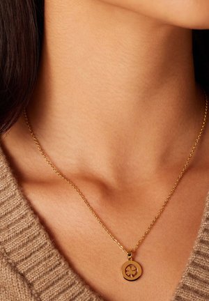Gouden ketting met een ronde hanger met een klavertje vier ontwerp. De ketting is delicaat en past mooi bij een gebreide, tan kleurige trui.