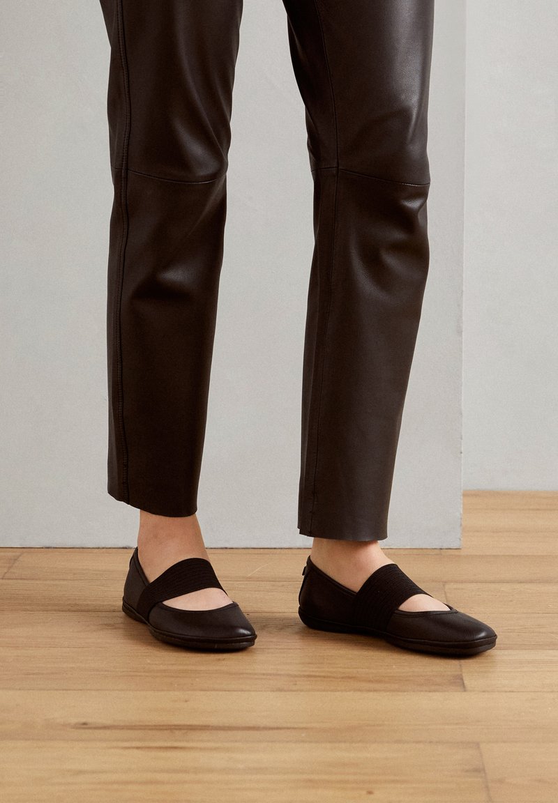 Zwarte ballet flats met een ronde neus, elastische band over de wreef en een gladde leren textuur. Draagt met zwarte aangesloten pantalons op een houten vloer.