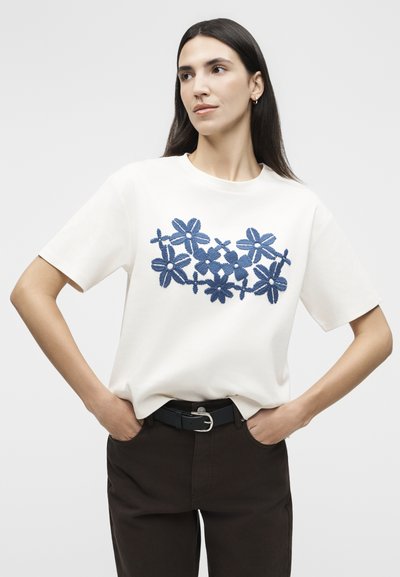 Γυναίκα με μακριά σκουρόχρωμα μαλλιά, φοράει λευκό πουκάμισο με κοντά μανίκια και μπλε λουλουδάτη κεντητική, μαύρο παντελόνι, τα χέρια στις τσέπες, κοιτάζει προς τα πλάγια.