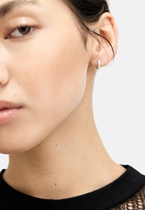 Silver hoops med små cirkulära detaljer, bärs i örat. Modellen har ljus hy och mörkt hår, klädd i en svart texturerad topp.