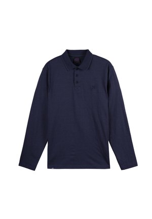 Jeune homme aux cheveux bouclés porte un polo à manches longues bleu marine, un pantalon gris et des baskets noires avec des lacets blancs, debout devant un fond uni.