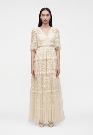 Femme portant une longue robe en dentelle crème avec broderie florale, manches bouffantes mi-longues et col en V, debout devant un fond blanc uni.