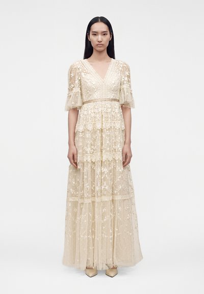 Femme portant une longue robe en dentelle crème avec broderie florale, manches bouffantes mi-longues et col en V, debout devant un fond blanc uni.