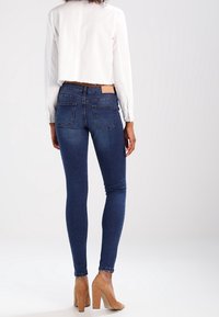 Jeans skinny in denim blu con vita alta, caratterizzati da un design della tasca posteriore e un'etichetta in pelle marrone chiaro sulla cintura. Abbinato a un top bianco cropped.