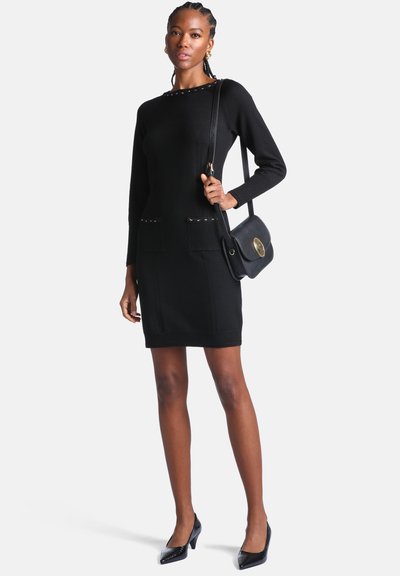 Femme portant une robe en maille noire à manches longues avec des détails à clous, des talons noirs et un sac à épaule noir avec une fermeture dorée, debout devant un fond blanc.