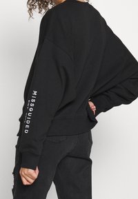 Sweat-shirt noir surdimensionné avec épaules tombantes, poignets et ourlet côtelés. Dispose d'un logo texte blanc sur la manche gauche. Texture de tissu lisse.