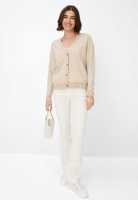 Beige- und cremefarbener gestreifter Cardigan mit V-Ausschnitt, Knopfleiste und gerippten Bündchen, kombiniert mit weißen Hosen und weißen Sneakers. Hält eine helle Tasche.