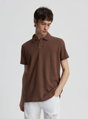 Giovane uomo con una polo marrone a maniche corte con trama e jeans bianchi strappati, guarda verso il basso con una mano in tasca.