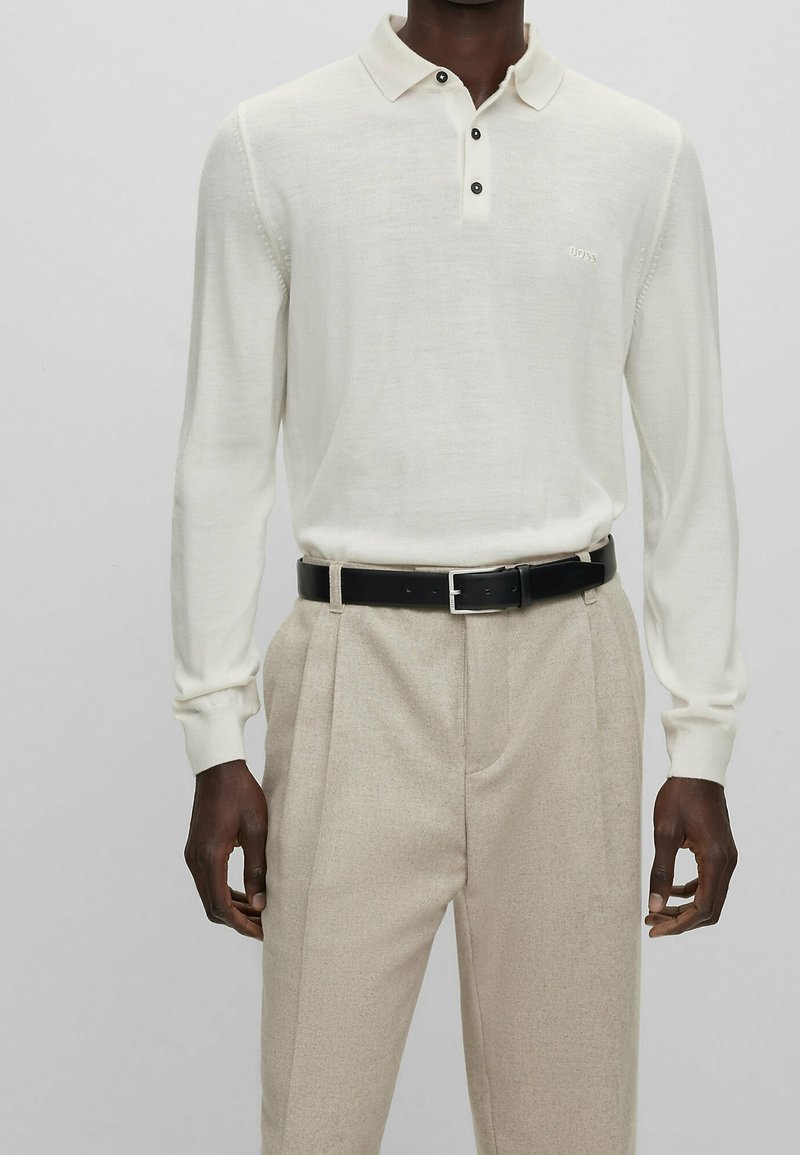 Polo blanc à manches longues avec trois boutons noirs et un logo BOSS discret. Associé à un pantalon beige clair sur mesure et une ceinture noire.