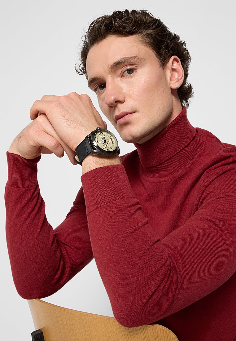 Reloj negro con una esfera grande y acentos plateados en una correa negra, llevado en una muñeca, con un modelo que lleva un suéter de cuello alto rojo ajustado.