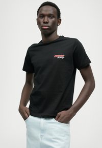 Svart kortermet t-skjorte med en rød og hvit logo på venstre bryst. Stoffet er glatt, og den har et klassisk rund hals-design.