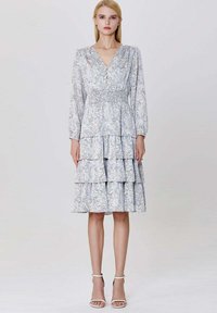 Femme portant une robe fleurie à col en V léger avec des manches longues, une jupe à volants et des talons à brides blanches, se tenant devant un fond uni.