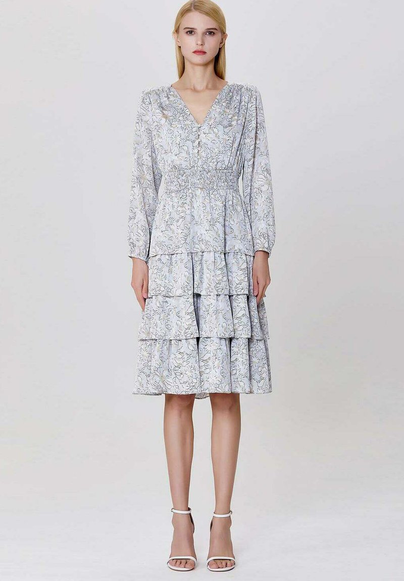 Femme portant une robe fleurie à col en V léger avec des manches longues, une jupe à volants et des talons à brides blanches, se tenant devant un fond uni.