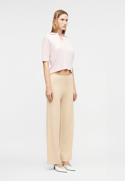 Γυναίκα που στέκεται, φοράει ανοιχτό ροζ μπλουζάκι με κοντά μανίκια και γιακά, μπεζ παντελόνι φαρδύ στο κάτω μέρος και λευκά παπούτσια με τακούνι, με φόντο απλό ανοιχτό γκρι.