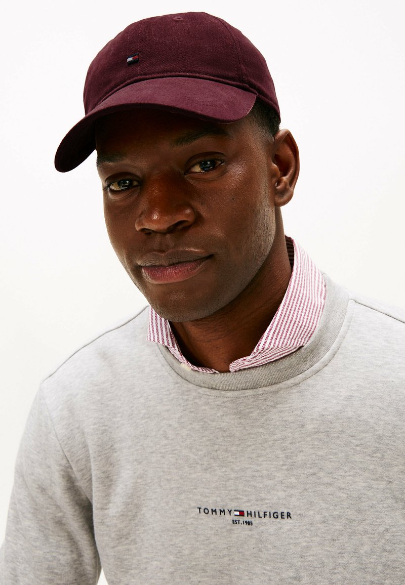 Maroon baseball cap with small logo, paired with a gray sweatshirt featuring a embroidered brand label, and a striped collar visible underneath.