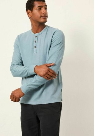 Light blue long-sleeve henley shirt with three button placket, textured fabric, ribbed cuffs, and side slits, paired with dark pants.