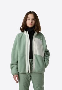 Jachetă din fleece verde cu accente crem, închidere cu fermoar și patch logo. Purtată peste un pulover negru cu guler înalt, combinată cu pantaloni verde deschis.