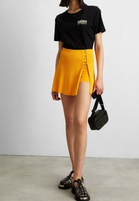 T-shirt noir à manches courtes avec texte, assorti à une mini-jupe plissée jaune vif avec boutons sur le côté. Sac bandoulière noir et sandales à plateforme.