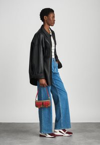 Μαύρη δερμάτινη oversized ζακέτα, ριγέ μπλούζα, ψηλόμεσες μπλε φαρδιές τζιν, κόκκινη τσάντα με σχέδια, μπορντό και λευκά αθλητικά παπούτσια.