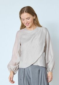 Lysegrå langærmet bluse i tekstureret stof med et asymmetrisk draperet design og opsamlede manchetter. Kombineret med et gråt skørt.