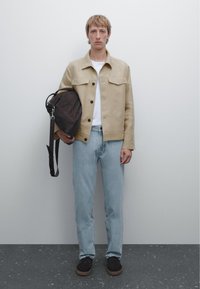 Beige mockajacka med knappstängning, vit t-shirt, ljusblå jeans och mörkbrun väska. Svarta skor med strukturerad sula. Neutral bakgrund.