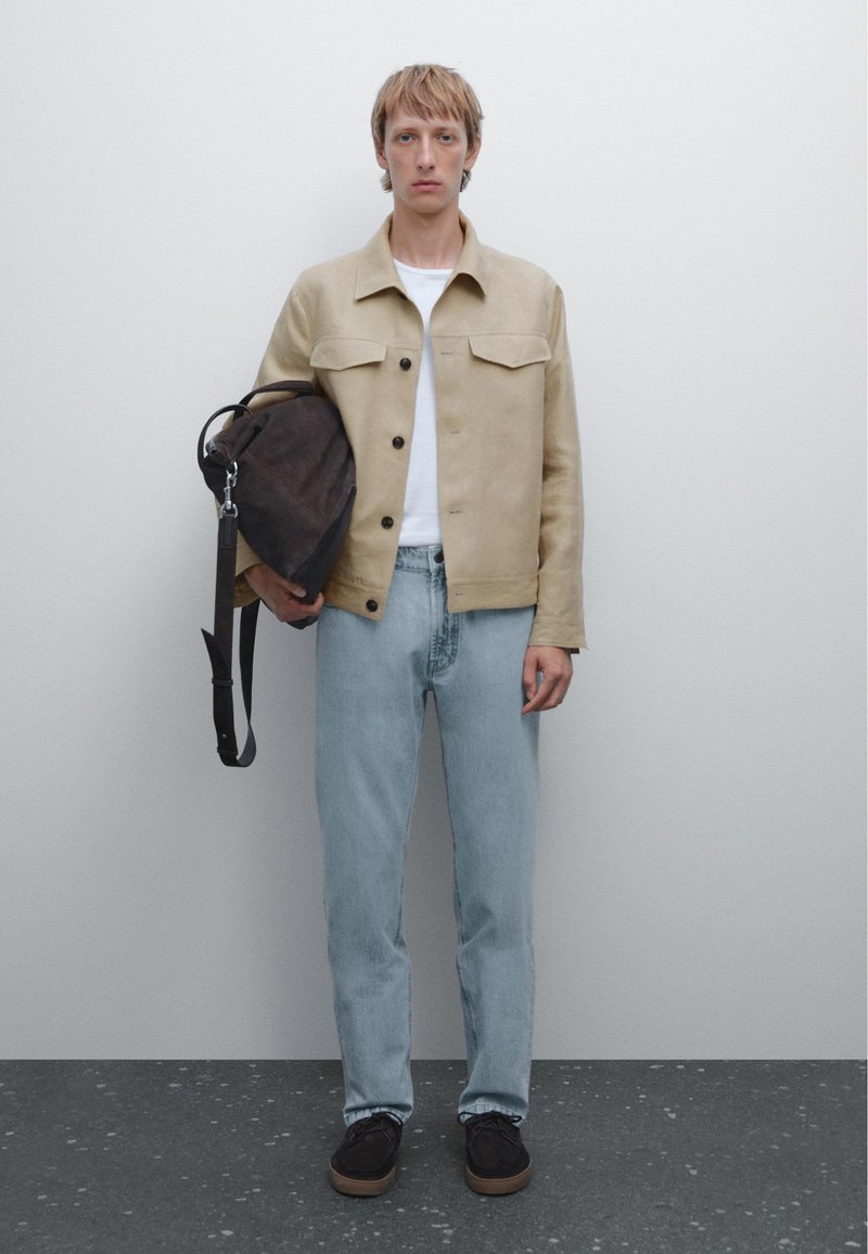 Beige mockajacka med knappstängning, vit t-shirt, ljusblå jeans och mörkbrun väska. Svarta skor med strukturerad sula. Neutral bakgrund.