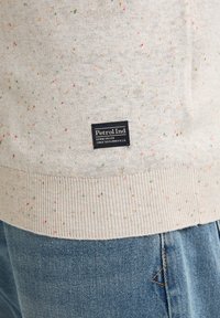 Lichtgrijze trui met veelkleurige spikkels, geribbelde zoom en een zwart "Petrol Ind" label; gecombineerd met blauwe denim jeans.