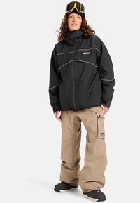 Zwart waterdicht jack met goudkleurige stiksels en hoge kraag, gecombineerd met losse beige cargobroeken en zwarte sneeuwlaarzen. Skibril op het voorhoofd.