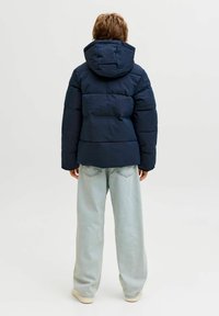 Person ar īsiem matiem, tērpies tumši zilā puffer jakā ar kapuci un gaiši zilos plašos džinsos, stāv ar muguru pret vienkāršu fonu.