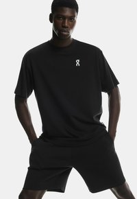 Tricou sport negru, cu mâneci scurte și decolteu rotund, având un mic logo alb pe piept, asortat cu pantaloni scurți negri.