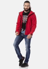 Rote Softshell-Jacke mit Kapuze, schwarzes Grafik-T-Shirt, blaue Slim-Fit-Jeans und schwarze Sneaker. Verfügt über Reißverschlusstaschen und kontrastierende Akzente.