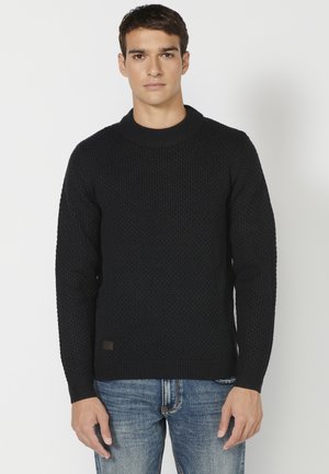Zwarte gebreide trui met een ronde halslijn, lange mouwen en een bruine logopatch aan de onderzijde van de voorkant, gedragen met blauwe jeans.