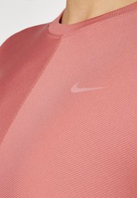 Rózsaszín sporting póló bordázott textúrával, kerek nyakkivágással és finom kontrasztos varrással. Kis fehér Nike logó található a mellkasán.