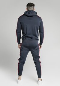 Marinblå träningsdräkt med röda sidosträck, tillverkad av slät tyg. Hoodien har dragsko. Joggingbyxorna har ribbade muddar och en figurnära design.