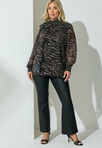 Schwarze und braune durchsichtige Bluse mit einem Zebra-Stripe-Muster und einem Schleifenkragen, kombiniert mit hochgeschnittenen schwarzen Kunstlederhosen und hohen Schuhen.
