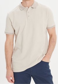 Beige polo tričko s texturovaným límcem a obšíváním rukávů, se třemi knoflíky a krátkými rukávy. Nosené s tmavými kalhotami.