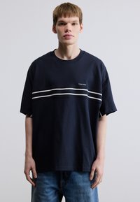 Μπλε ναυτικό βαμβακερό t-shirt με κοντά μανίκια, που διαθέτει μια λευκή οριζόντια ρίγα και ένα μικρό λογότυπο στο  στήθος. Χαλαρή εφαρμογή.